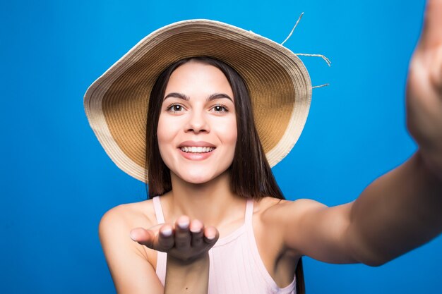 Young fashionable attractive girl in sunglasses and straw hat sends air kiss to camera. Nice playful and happy woman with red lips on blue wall. with copy space.