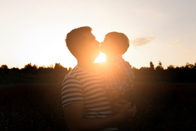 Young father kisses his toddler son on spring flower field at sunset.