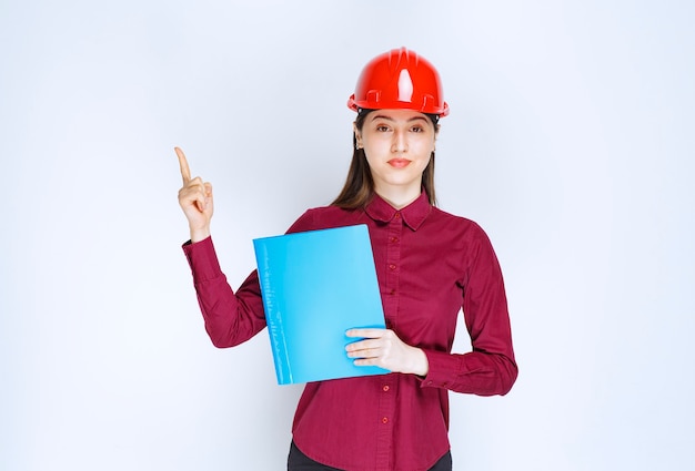 Young female architect in red helmet holding documents and pointing on white background. 