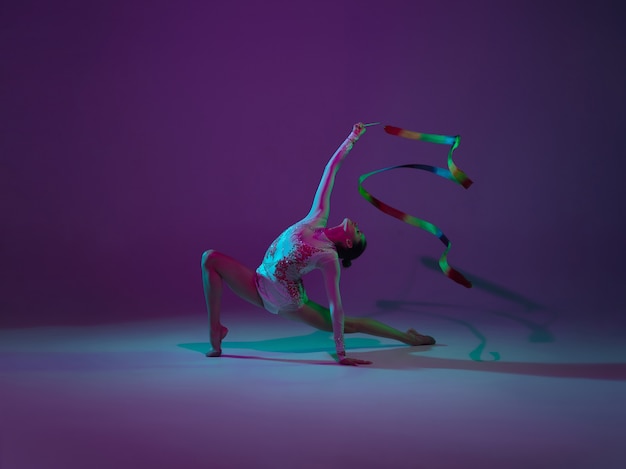 Young female athlete, rhythmic gymnastics artist dancing, training isolated on purple studio background with neon light. Beautiful girl practicing with equipment. Grace in performance.