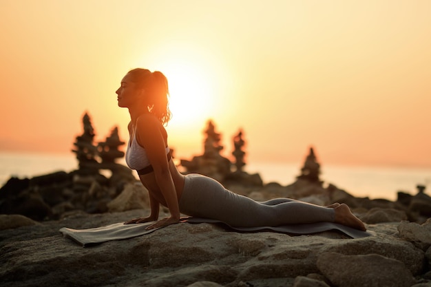 Young female athlete stretching while working out on rocky beach at sunset. Copy space.