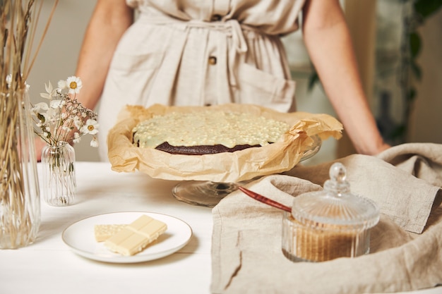 Free photo young female baker making a delicious chocolate cake with cream on a white table