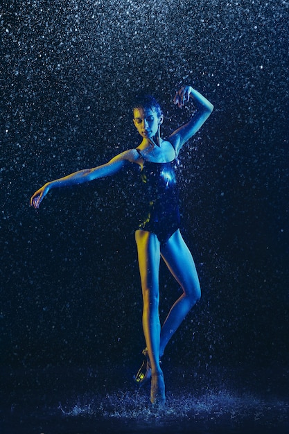 Young female ballet dancer performing under water drops and spray. Caucasian model dancing in neon lights. Attractive woman. Ballet and contemporary choreography concept. Creative art photo.