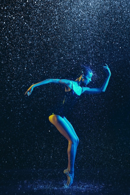 Young female ballet dancer performing under water drops and spray. Caucasian model dancing in neon lights. Attractive woman. Ballet and contemporary choreography concept. Creative art photo.