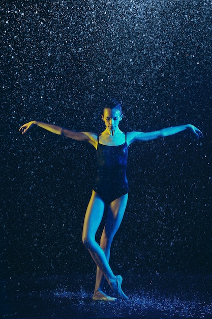 Young female ballet dancer performing under water drops and spray. Caucasian model dancing in neon lights. Attractive woman. Ballet and contemporary choreography concept. 