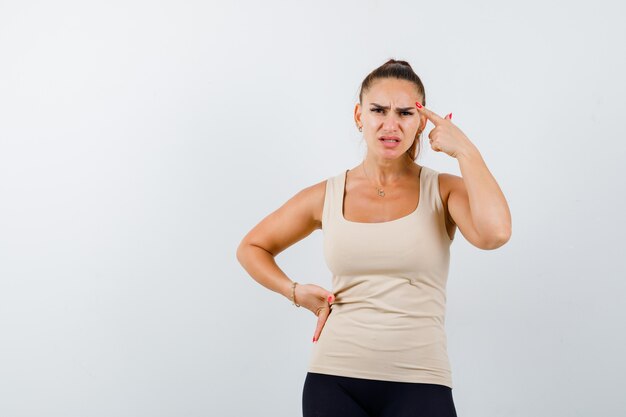 Young female in beige tank top standing in thinking pose while keeping hand on hip and looking pensive