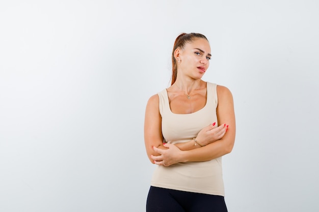 Young female in beige tank top standing with crossed arms while sticking out tongue and looking pretty