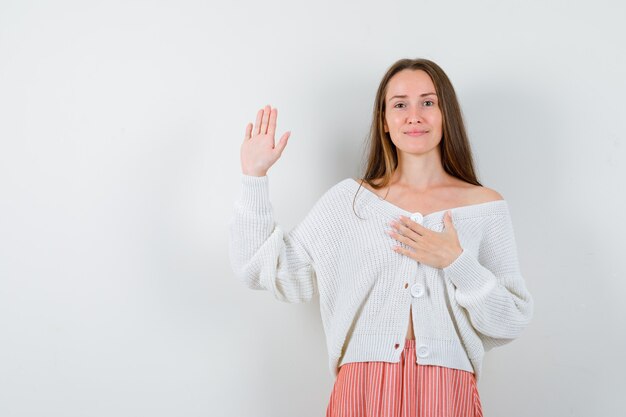 Young female in cardigan and skirt keeping hand on chest, showing palm isolated