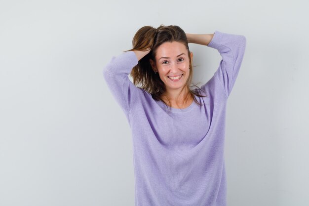 Young female clasping her hair with hands in lilac blouse and looking jolly. front view.