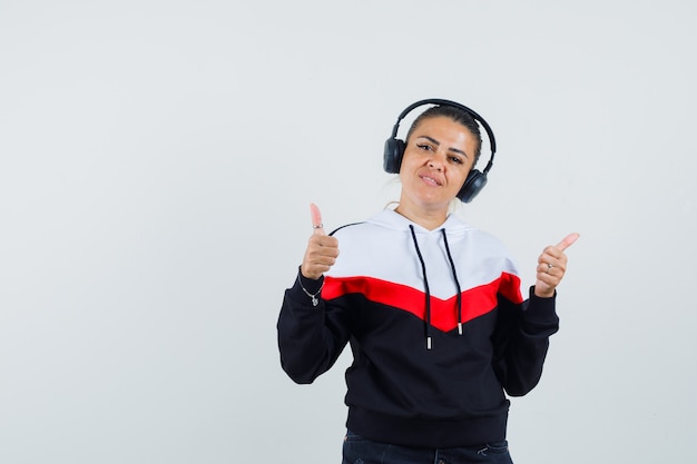 Free photo young female in colorful sweatshirt,earphones showing thumb up while listening music and looking pleased , front view.