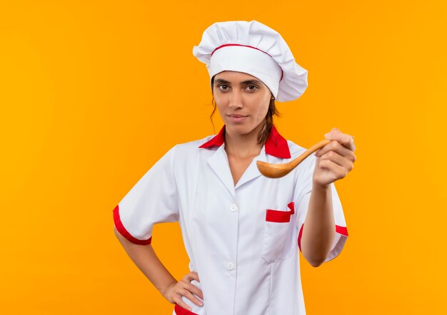 young female cook wearing chef uniform holding spoon on isolated yellow wall with copy space