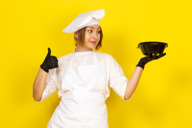 Free photo young female cooking in white cook suit and white cap in black gloves showing black bowl