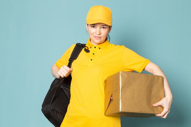 young female courier in yellow t-shirt yellow cap holding box on the blue wall