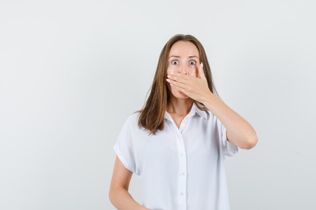 Young female covering her mouth with her hand in white blouse and looking shocked.