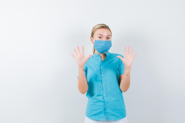 Young female doctor in medical uniform, mask showing palms in surrender gesture