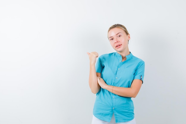 Young female doctor pointing back with thumb in medical uniform, mask and looking pretty