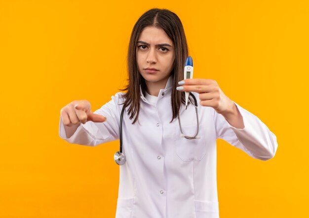 Young female doctor in white coat with stethoscope around her neck holding digital thermometer pointing with index finger to the front with serious face standing over orange wall