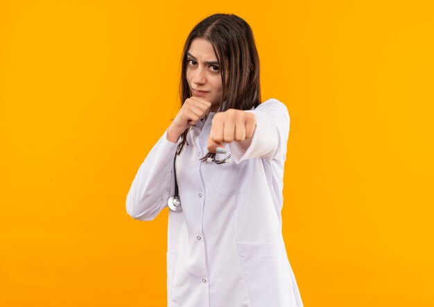 Young female doctor in white coat with stethoscope around her neck looking to the front with serious face posing like a boxer standing over orange wall