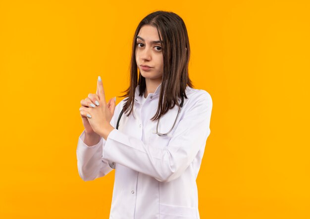 Young female doctor in white coat with stethoscope around her neck making pistol or gun gesture with fingers looking to the front with serious face standing over orange wall