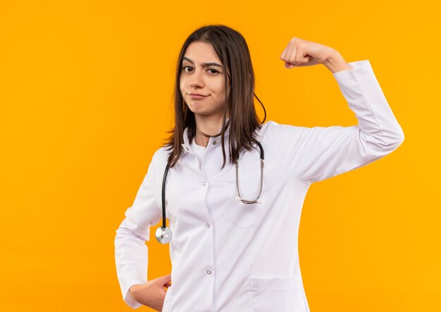 Young female doctor in white coat with stethoscope around her neck raising fist looking to the front with skeptic expression standing over orange wall