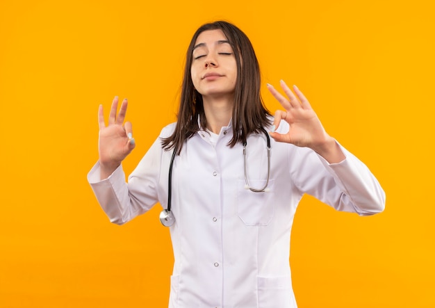 Young female doctor in white coat with stethoscope around her neck relaxing with closed eyes making meditation gesture with fingers standing over orange wall