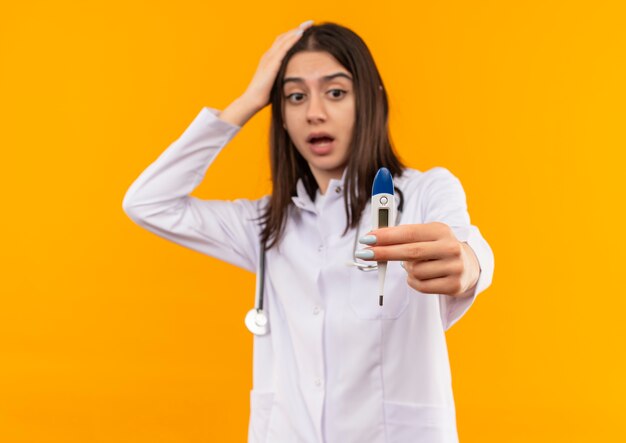 Young female doctor in white coat with stethoscope around her neck showing digital thermometer looking at it surprised standing over orange wall