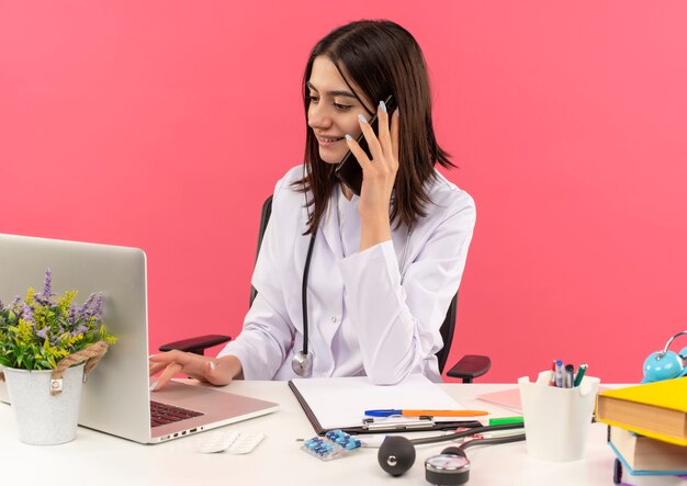 Free Photo young female doctor in white coat with stethoscope around her neck working on laptop computer and talking on mobile phone with smile on face sitting at the table over pink wall