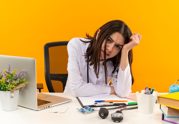 Young female doctor in white coat with stethoscope looking to the front confused and very anxious sitting at the table with laptop and documents over orange wall