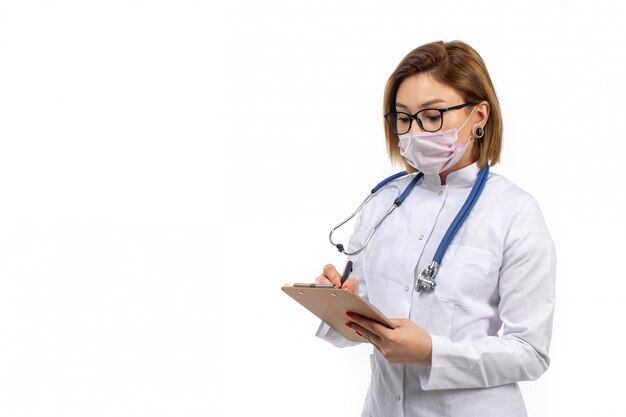 young female doctor in white medical suit with stethoscope in white protective mask writing down notes on the white