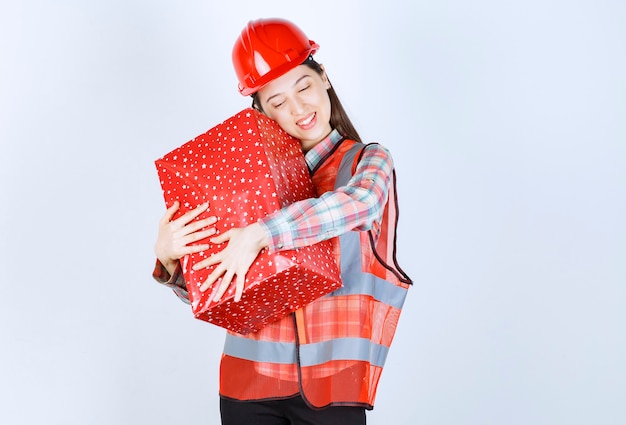 Young female engineer in red helmet hugs gift box. 