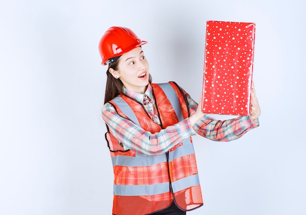 Young female engineer in red helmet looking at red gift box. 