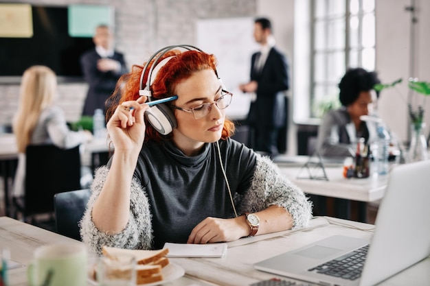 Young female entrepreneur using laptop and thinking about email she has received while listening music on headphones at work There are people in the background