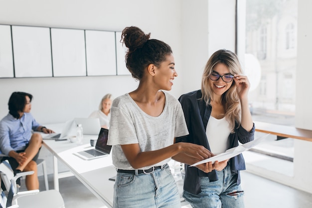 Young female executive explaines new strategy to blonde employee in glasses and smiling. Indoor portrait of multicultural collective working on project in office and using laptop.