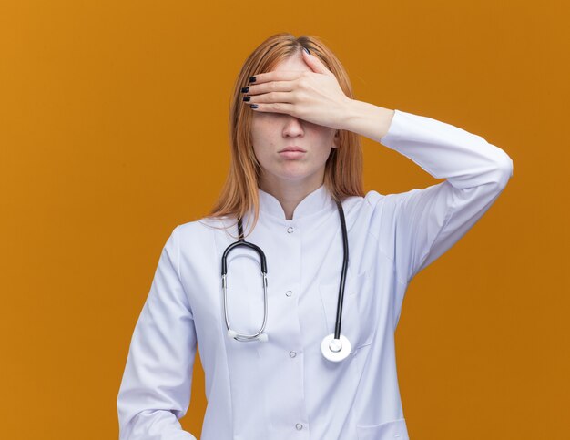 Young female ginger doctor wearing medical robe and stethoscope covering eyes with hand isolated on orange wall