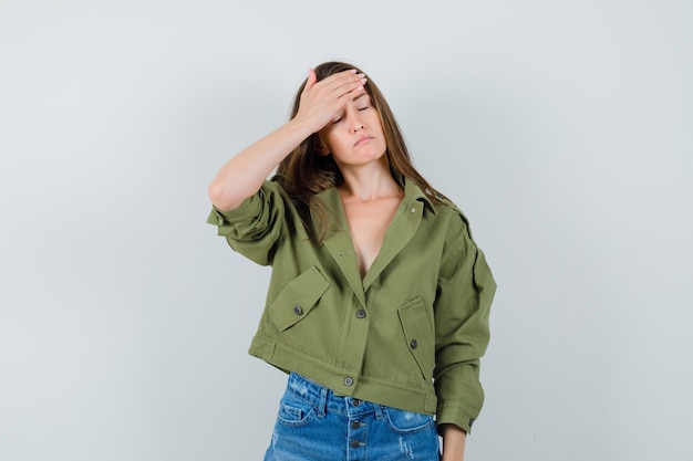 Young female in green jacket, shorts holding hand on forehead and looking tired , front view.