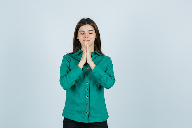 Young female in green shirt holding hands in praying gesture and looking hopeful , front view.