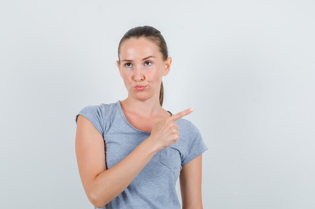 Young female in grey t-shirt pointing to side and looking pensive , front view.