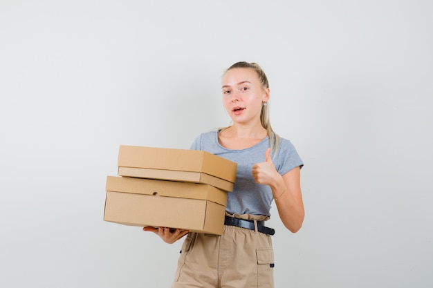 Young female holding cardboard boxes, showing thumb up in t-shirt, pants and looking cheery. front view.