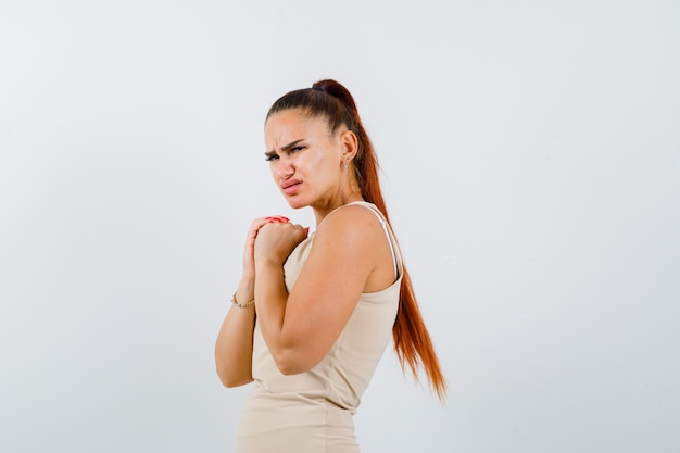 Young female holding clasped hands on chest in beige tank top and looking displeased , front view.