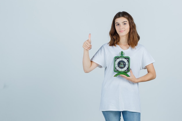 Free photo young female holding clock, showing thumb up in white t-shirt, jeans and looking cheery. front view.