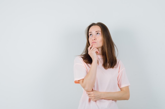 Young female holding finger on chin in pink t-shirt and looking indecisive. front view.