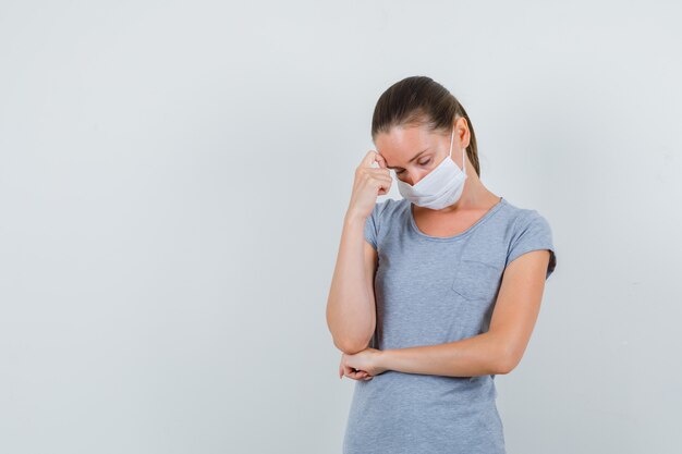 Young female holding finger on forehead in grey t-shirt, mask and looking exhausted. front view.