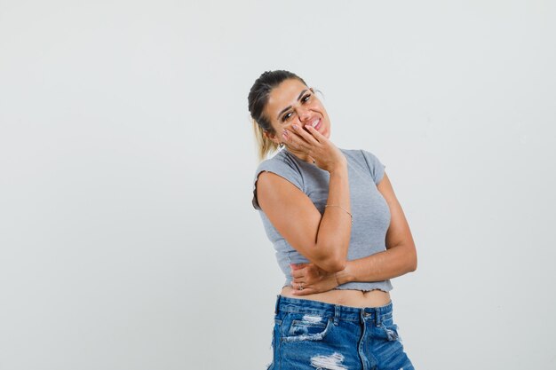 Young female holding hand on cheek in t-shirt, shorts and looking glad