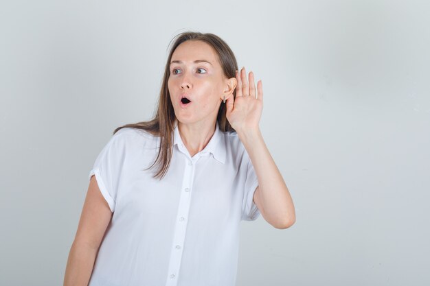 Young female holding hand to ear in white shirt and looking surprised