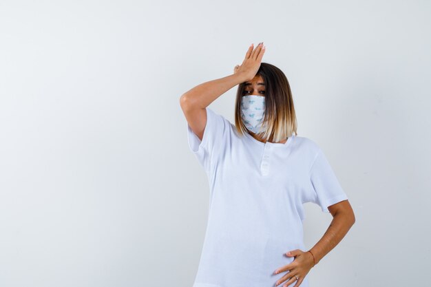 Young female holding hand on forehead in t-shirt, mask and looking forgetful , front view.