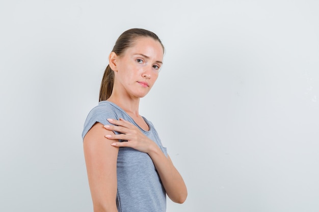 Young female holding hand near chest in grey t-shirt and looking serious. front view.