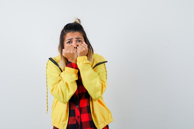 Young female holding hands on face in checkered shirt, jacket and looking scared , front view.