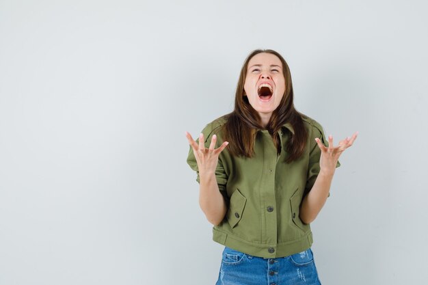 Young female in jacket, shorts raising hands in aggressive manner while screaming , front view.