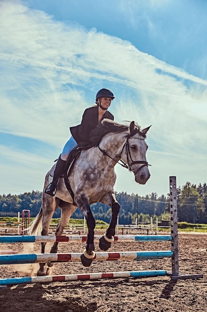 Young female jockey on dapple gray horse jumping over hurdle in the open arena.