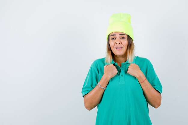 Young female keeping clenched fists on chest in polo t-shirt, beanie and looking grumpy , front view.